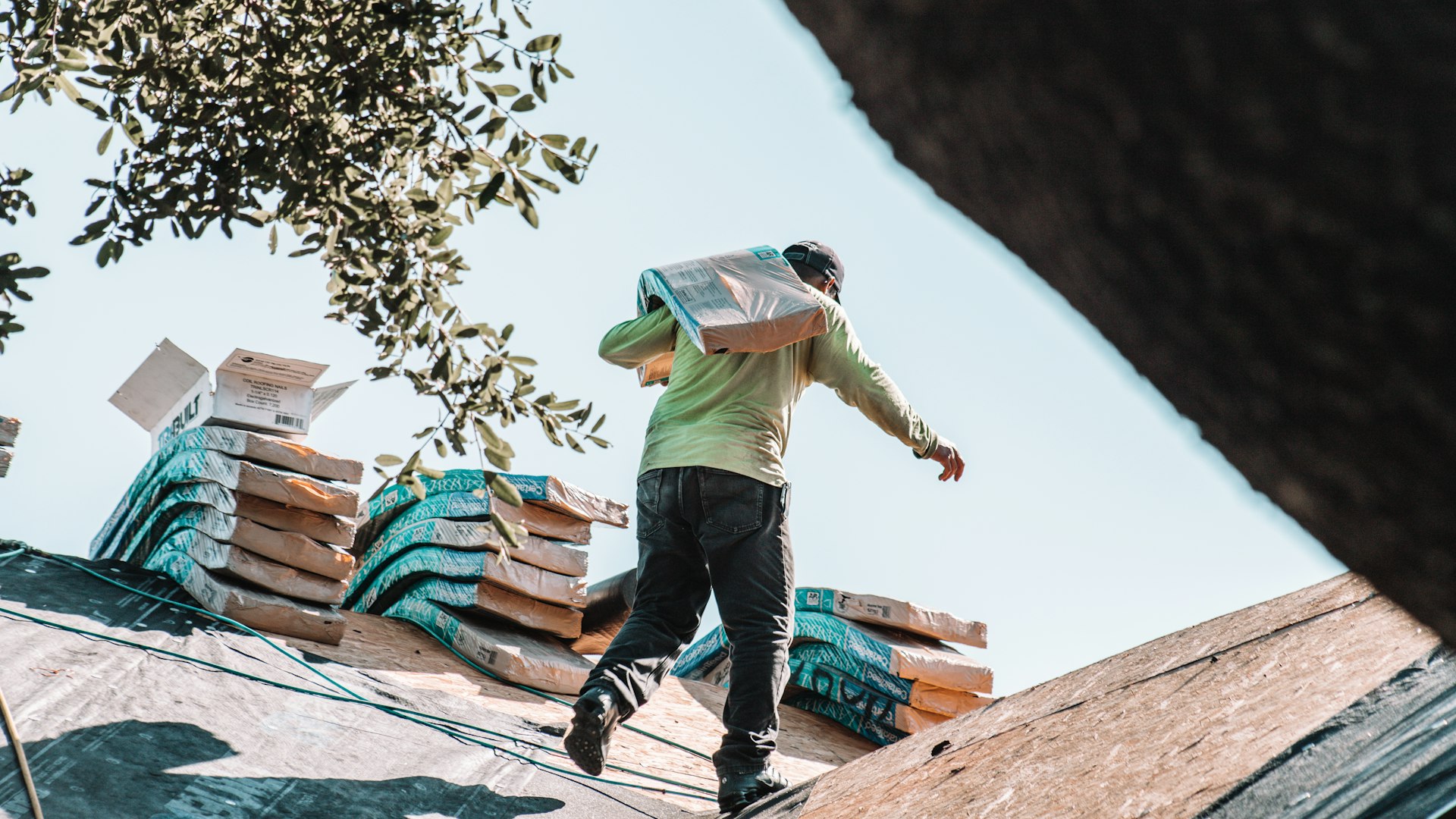 A roofer carrying a bundle of shingles across a residential roof
